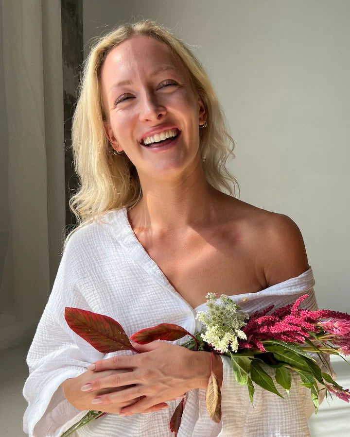 A woman with long blonde hair smiles brightly at the camera, wearing a white top draped over one shoulder. She holds a colorful bouquet of flowers, including red and white blooms, standing in front of a plain background.